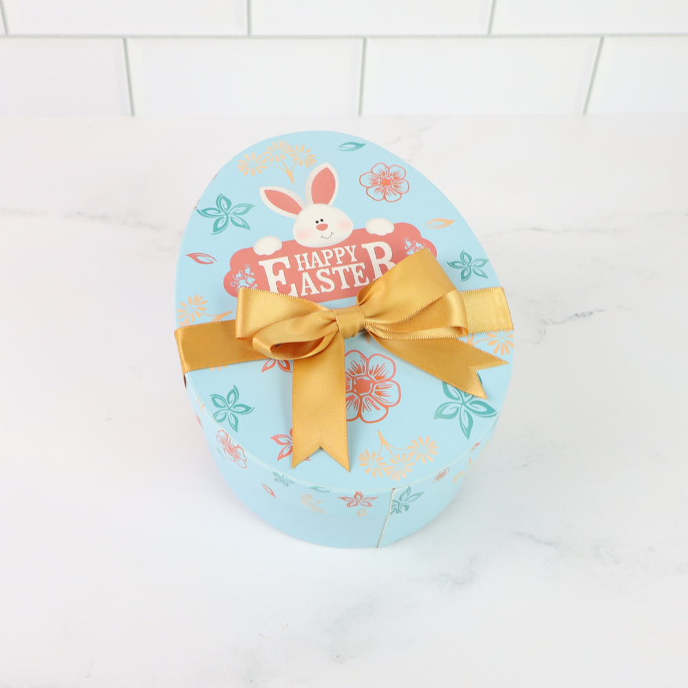 Easter egg-shaped gift box with a gold bow on a white tiled background