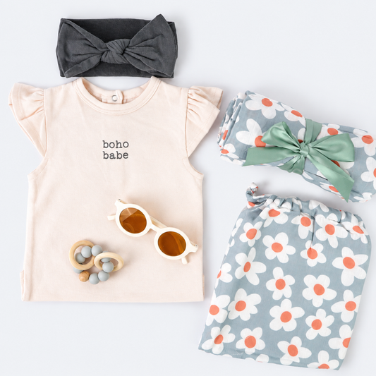 A baby outfit flat lay featuring a light peach short-sleeve top with 'boho babe' printed on the chest, a dark gray bow headband, a sage green ribbon-wrapped blanket, and matching sage green and white daisy-patterned shorts. A wooden teething toy with amber-colored sunglasses is also included, all arranged on a clean white background.
