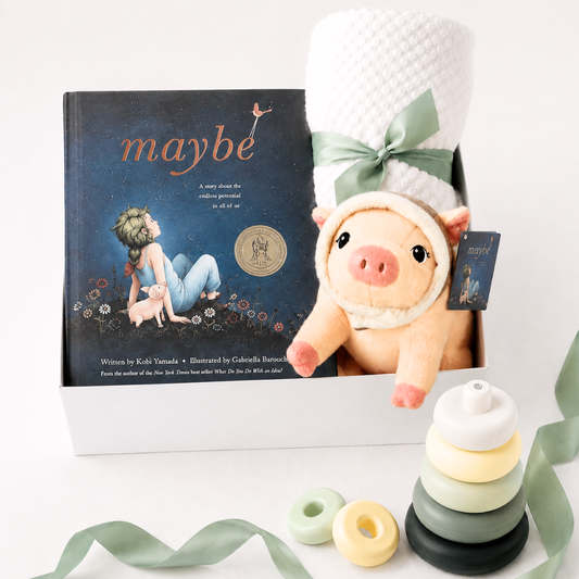 A curated gift set featuring the children's book 'maybe' by Kobi Yamada, a plush pig toy wearing a white knit hat with a sage green ribbon, a white textured blanket, and a wooden stacking ring toy, all arranged in a white box on a white background.
