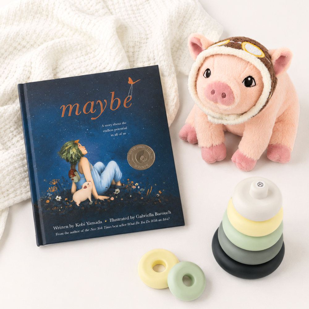 A children's book titled "maybe" is styled on a white surface with a cream knit blanket, a pink plush pig wearing a brown aviator hat, a stack of soft stacking rings in white, pale yellow, pale green, and black, and two small ring toys in pale yellow and pale green.