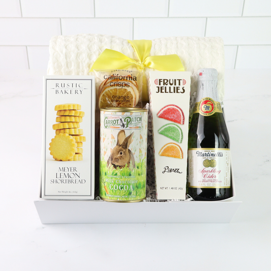 Gift basket with various items including a bottle of sparkling cider, cookies, and a can of carrots on a white background.