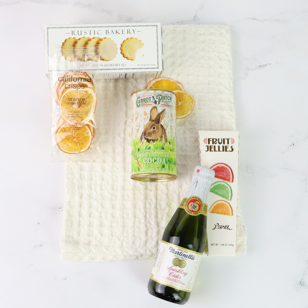 Assorted items including a bottle of sparkling wine, a can of carrot cake, and a package of orange slices on a textured white surface.