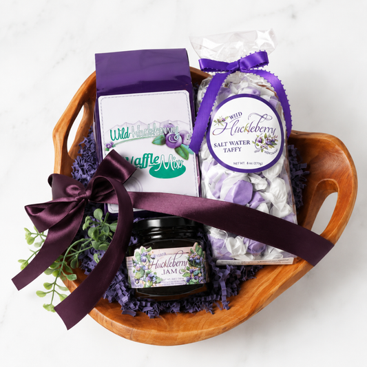 A huckleberry-themed gift basket arranged in a wooden bowl, featuring a bag of Wild Huckberry Waffle Mix, a bag of Wild Huckberry Salt Water Taffy, a jar of Wild Huckberry Jam, and a purple ribbon, set on a white marble background.