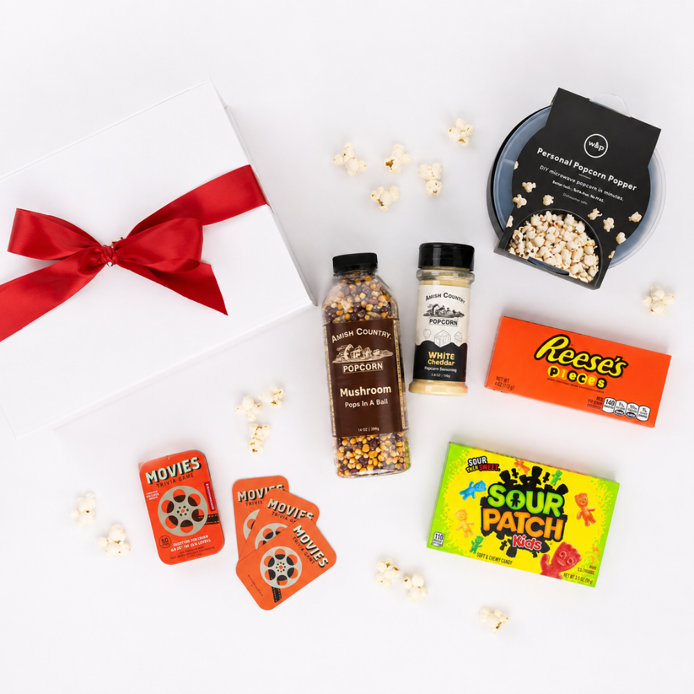 A curated gift box flat lay featuring a white box with a red ribbon, a jar of Raw Country Popcorn, a bottle of White Pepper, a bag of Personal Poppin' Popcorn, a box of Reese's Pieces, a pack of Sour Patch Kids, and movie ticket inserts on a white background with scattered popcorn.