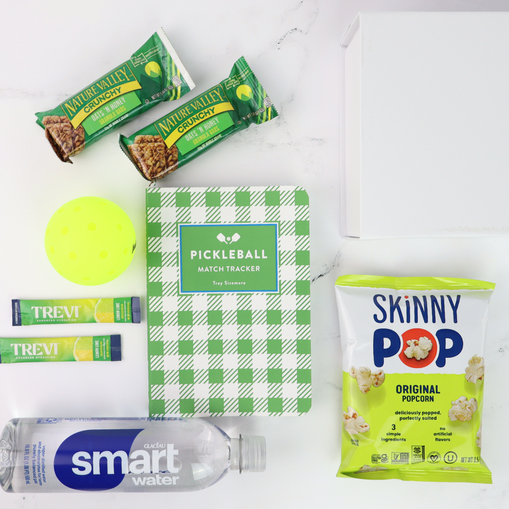 pickleball gift set with a Collection of snacks and a pickleball match tracker on a white surface