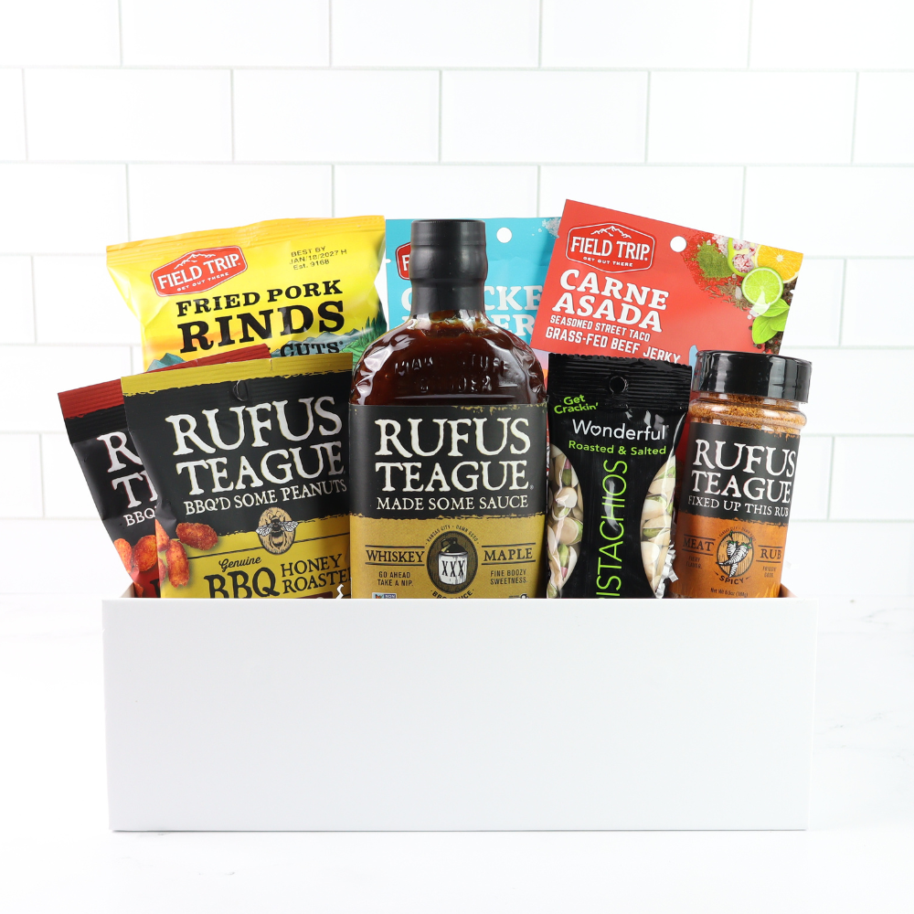 Assorted food products including a bottle of Rufus Teague whiskey and various snack packages on a white background.