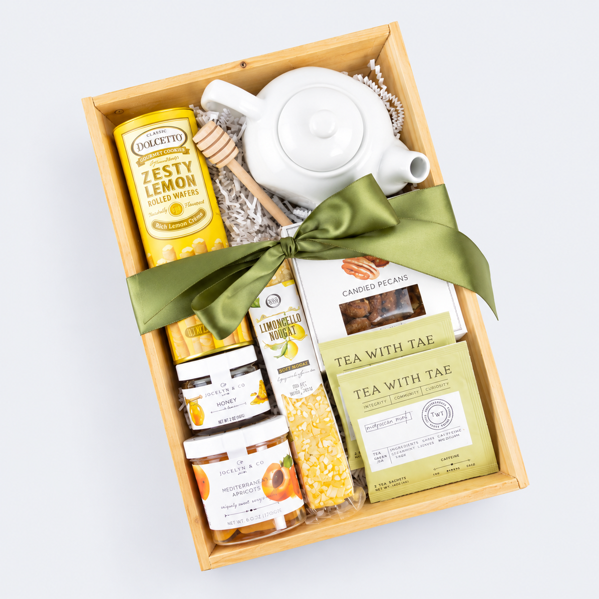 A wooden gift box filled with tea and snack items, tied with an olive green ribbon. Visible items include a white teapot, a yellow can of Dolce & Gabbana Zesty Lemon Infused Water, boxes of Tea with Tae, and jars of honey and candied pecans, all arranged on a light gray background.