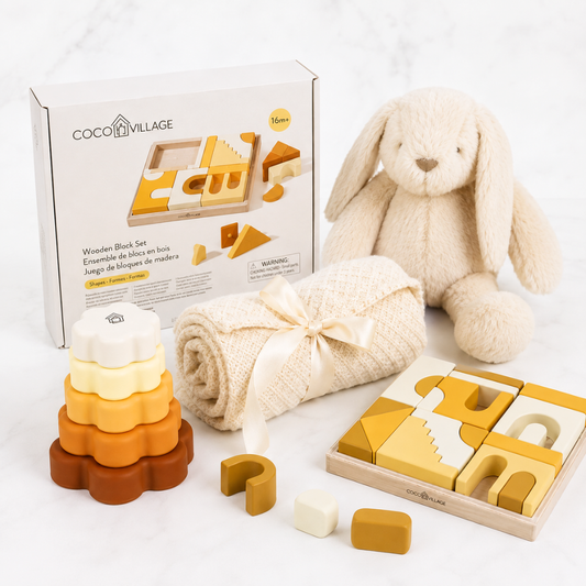 Coco Village wooden block set displayed with its packaging box, a plush bunny toy, a rolled cream blanket tied with a ribbon, a wooden stacking tower, a puzzle box, and loose blocks on a light marble surface.