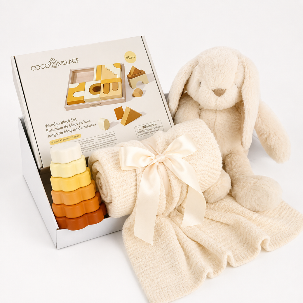 Coco Village wooden blocks set in a white display box, styled with a cream bunny plush toy, a cream waffle-knit blanket tied with a satin bow, and a small stack of wooden blocks in cream, yellow, and orange on a white background.